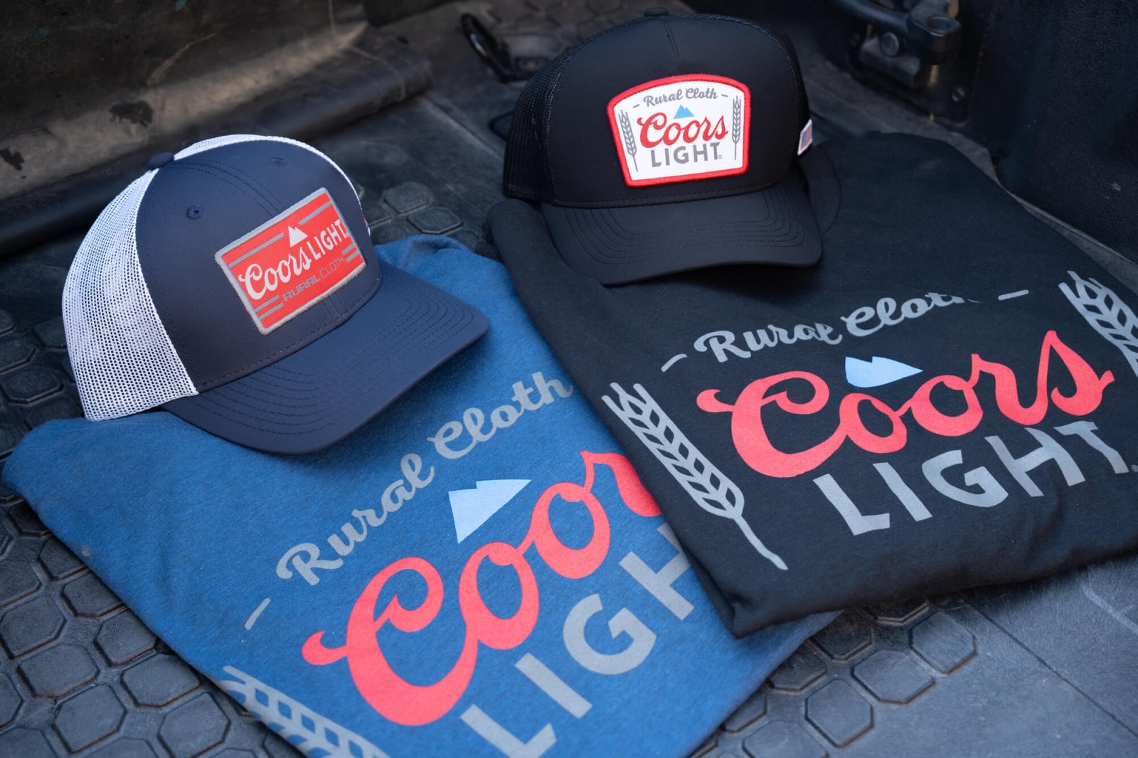Two t-shirts and two trucker hats with “Rural Cloth” and “Coors Light” logos are displayed on a textured surface, one shirt and hat in blue and the other shirt and hat in black.