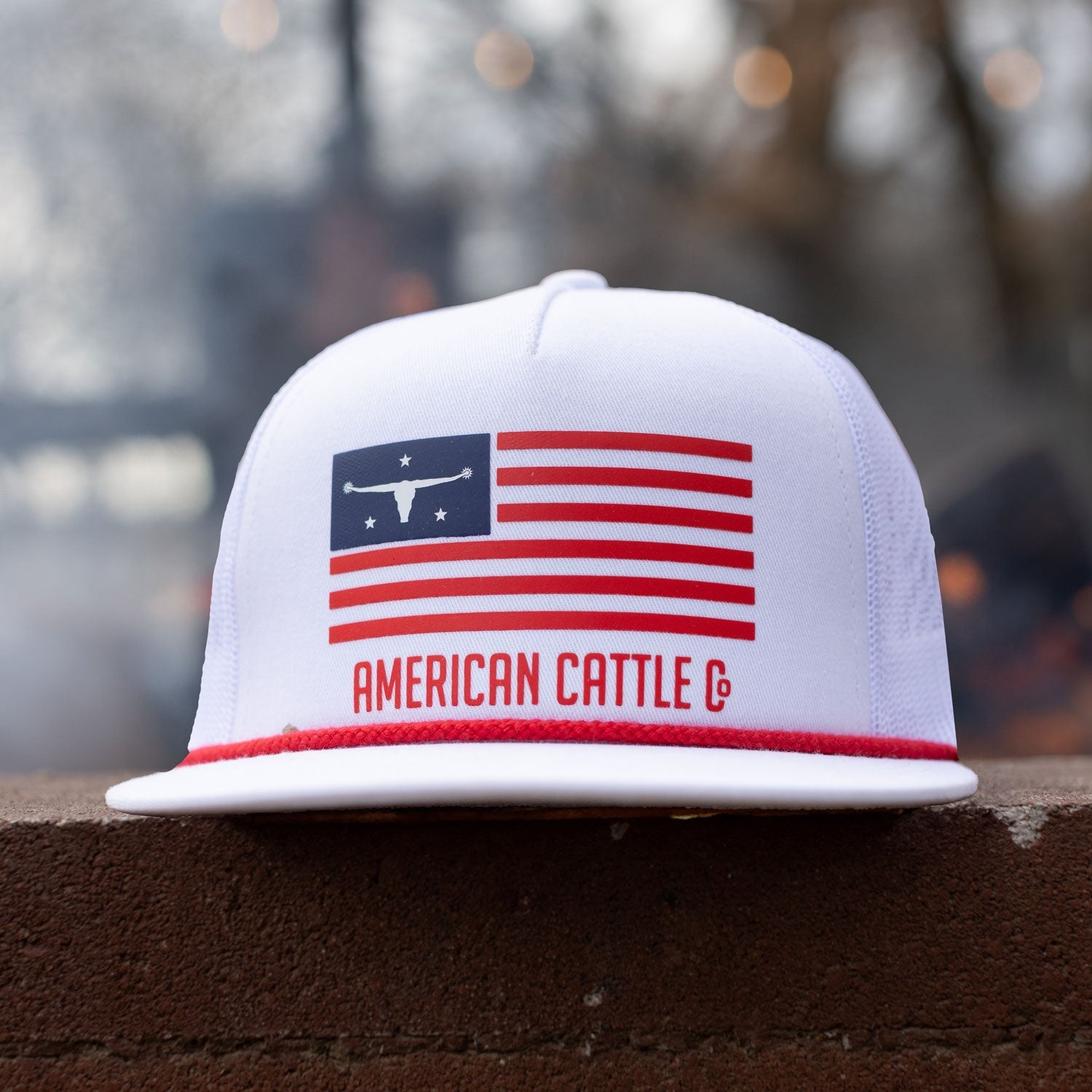 A man wearing sunglasses and a cap with "America" text is holding a Coors Light can while grilling outdoors. There is a U.S. flag and colorful flowers in the blurred background.