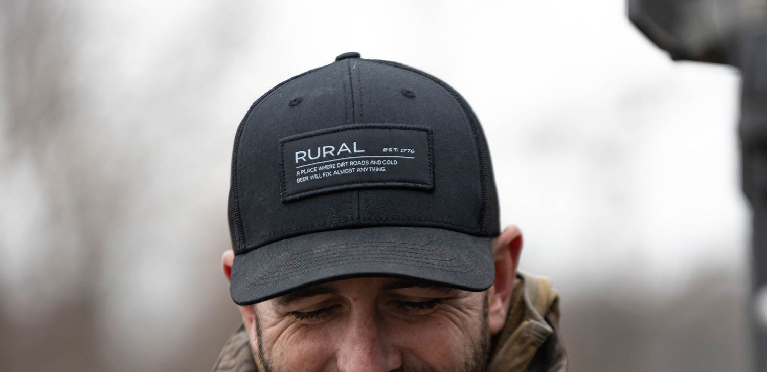 A man in an outdoor setting wears a black baseball cap with a patch that reads "RURAL" and additional smaller text. Only the upper part of his face is visible; he is looking down and smiling.