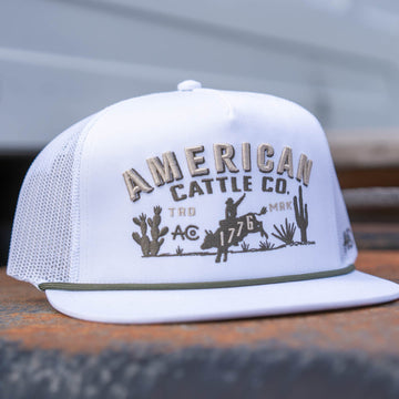 The American Cattle Co 8-Second Hat is a white mesh snapback with "AMERICAN CATTLE CO." embroidered on the front, showcasing a cowboy on horseback, two cacti, and bold decorative details in a flatbill profile.