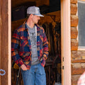 A man wearing the Rural Cloth Lumberjacket and a gray cap stands in the doorway of a wooden log cabin, one hand in his pocket, looking to his left.