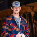 A man stands indoors looking forward, wearing a gray and blue trucker hat, a gray t-shirt, and the Rural Cloth Lumberjacket—a blue, red, and orange chore jacket that also functions as a warm work jacket.