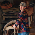 A man wearing the Rural Cloth Lumberjacket and a cap stands indoors, holding a saddle. Folded blankets and tack equipment are visible in the background as he looks neutrally at the camera.