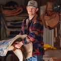 A man in a Rural Cloth Lumberjacket and baseball cap holds a saddle blanket and saddle indoors, with blankets and gear in the background.