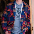 A person stands partially in a doorway with one hand in their pocket, wearing the Rural Cloth Lumberjacket men’s chore jacket in blue, red, and brown over a gray graphic t-shirt. The person's head is not fully visible.