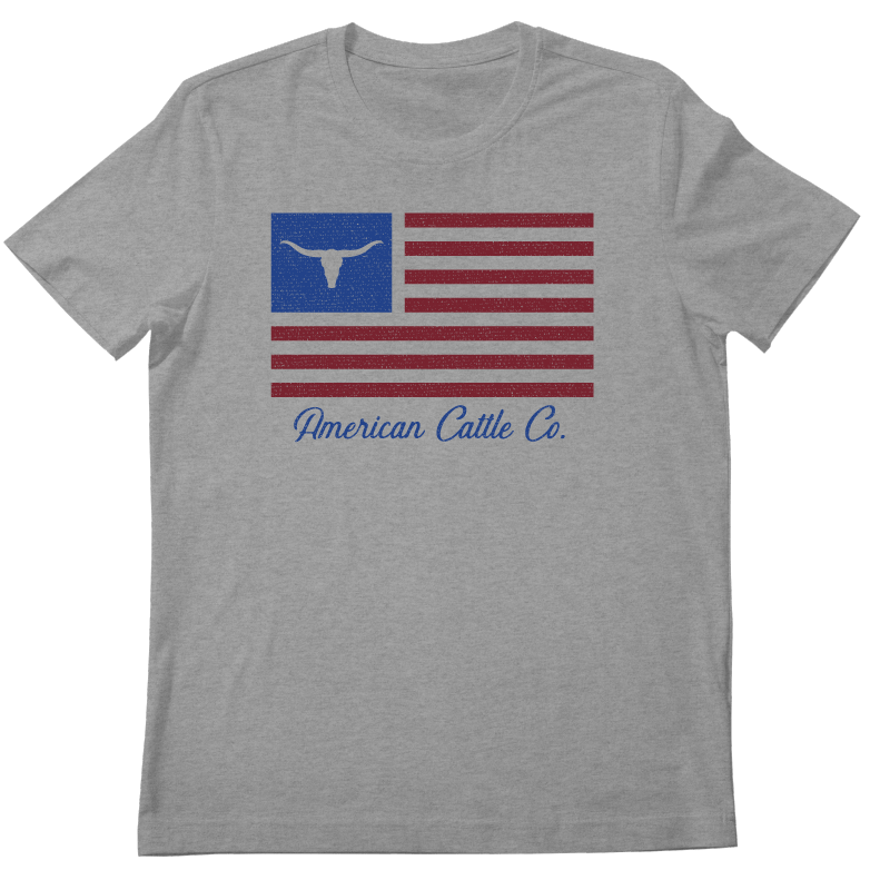 The Rural Cloth ACC Flag Tee is a gray patriotic shirt featuring a stylized American flag with a longhorn head in the blue field and "American Cattle Co." printed below in bold blue cursive.