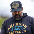 A bearded man looks down, wearing a Rural Cloth ACC Mark Pullover and cap, both featuring “American Cattle Co. 1776” in yellow and white, standing out against a blurred outdoor background.