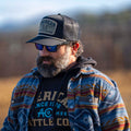 A bearded man wears sunglasses, a black patch cap, a striped jacket, and a Rural Cloth ACC Mark Pullover. He is outdoors with blurred natural scenery in the background.