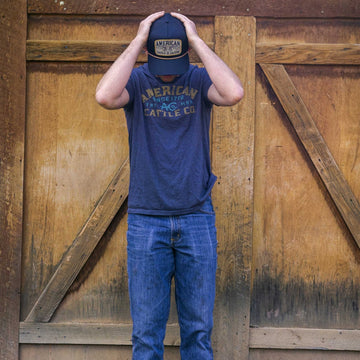 A person in blue jeans and an American Cattle Co ACC Mark Tee with a rib-knit neck stands before a wooden door, holding a matching cap in their hands. Their face is hidden from view.