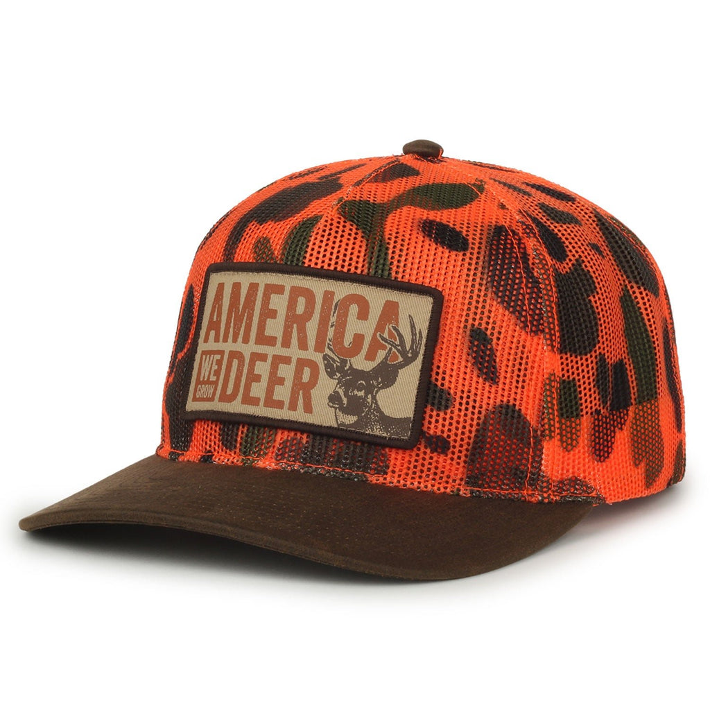 The American Cattle Co America We Grow Deer Hat features an orange and black blaze camo design, a brown brim, and an embroidered front patch with a deer head illustration and the text “AMERICA NEEDS DEER.”.