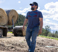 A man wearing sunglasses, a cap, jeans, and a blue Rural Cloth Billboard Tee walks outdoors by large hay bales on a sunny day with trees and mountains in the background.