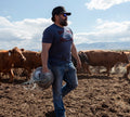 A man in a Rural Cloth Billboard Tee, sunglasses, and jeans carries hay through a dirt field among brown cows under a partly cloudy sky, capturing the rugged spirit of American ranch life.