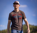 A man in the Rural Cloth Billboard Tee, blue jeans, and a white and red baseball cap strolls outdoors below a blue sky with green trees—a timeless rural look from the trusted brand.