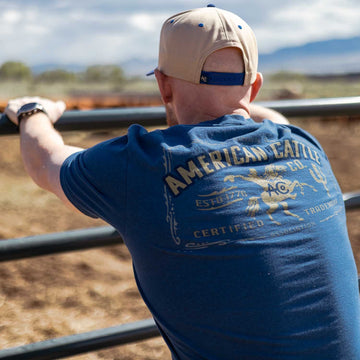 A person in a blue Bucking Horse Tee by American Cattle Co and a beige cap faces away, leaning on a metal fence and gazing at a cattle field. The tee features a cowboy graphic and retro rodeo text on the back.