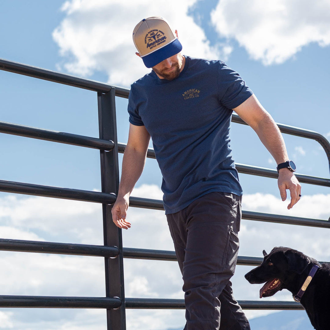 A man in an American Cattle Co Bucking Horse Tee, dark pants, and a baseball cap walks outdoors by a metal fence with a black dog under partly cloudy skies.