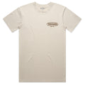 The Bucktown Caddie Tee by American Cattle Co is a plain beige short-sleeve shirt with a small oval brown and black geometric design on the left chest and a round neckline, creating a minimalist style with no other visible logos or patterns.