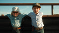 Two young children stand side by side outdoors at Bucktown XV Ranch in Stephenville, TX, both wearing Bucktown Outrider Hats by Rural Cloth. They rest their arms on a metal fence and look directly at the camera.