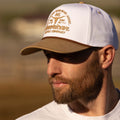 A man with a brown beard wears the Certified Ranchers Association Hat by Rural Cloth, a white and tan embroidered snapback. He looks to his left in a white shirt with a softly blurred background.