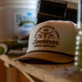 The Certified Ranchers Association Hat by Rural Cloth, a beige cap featuring front branding, sits on a wooden shelf with tools and containers in the background.