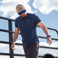 A man in a blue T-shirt, gray cargo shorts, and a Rural Cloth Certified Ranchers Association Hat walks outdoors near a black metal fence under a blue sky, with part of a black dog's head visible at the bottom right.