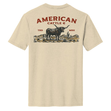The American Cattle Co "Desert Longhorn Tee" is a beige shirt featuring a longhorn cow, cacti, and plants graphic on the back with bold red “AMERICAN CATTLE CO.” text above and “TRD” & “MRK” at the sides—a true Western desert style.