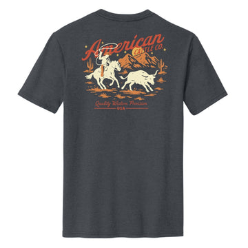 The Frontier Tee by American Cattle Co is a dark gray shirt with a vintage western vibe, featuring a cowboy herding cattle, mountain and cactus graphics, and bold red-orange "American Cattle Co." and "Quality Western Provision USA" text.