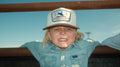 A child in a light denim shirt and a white Rural Cloth Last Out Hat western snapback leans against a metal railing, staring at the camera with a scrunched, determined look. A clear blue sky stretches in the background.