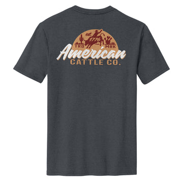 The Liberty Buck Tee by American Cattle Co is a charcoal western shirt with a bold back graphic of cowboys, cacti, and "est 1776 American Cattle Co." in tan, brown, and white—ideal for fans of the classic Liberty Buck style.