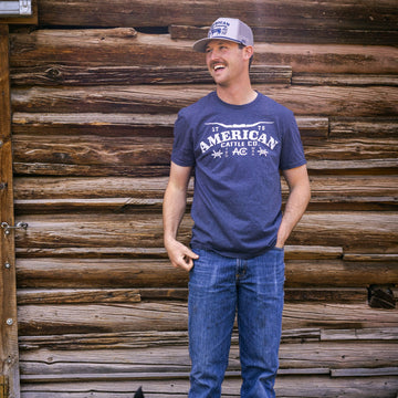 A man in a blue Longhorn Barb Tee by American Cattle Co, paired with blue jeans and a baseball cap, stands before a rustic wooden wall, exuding classic Western heritage style.