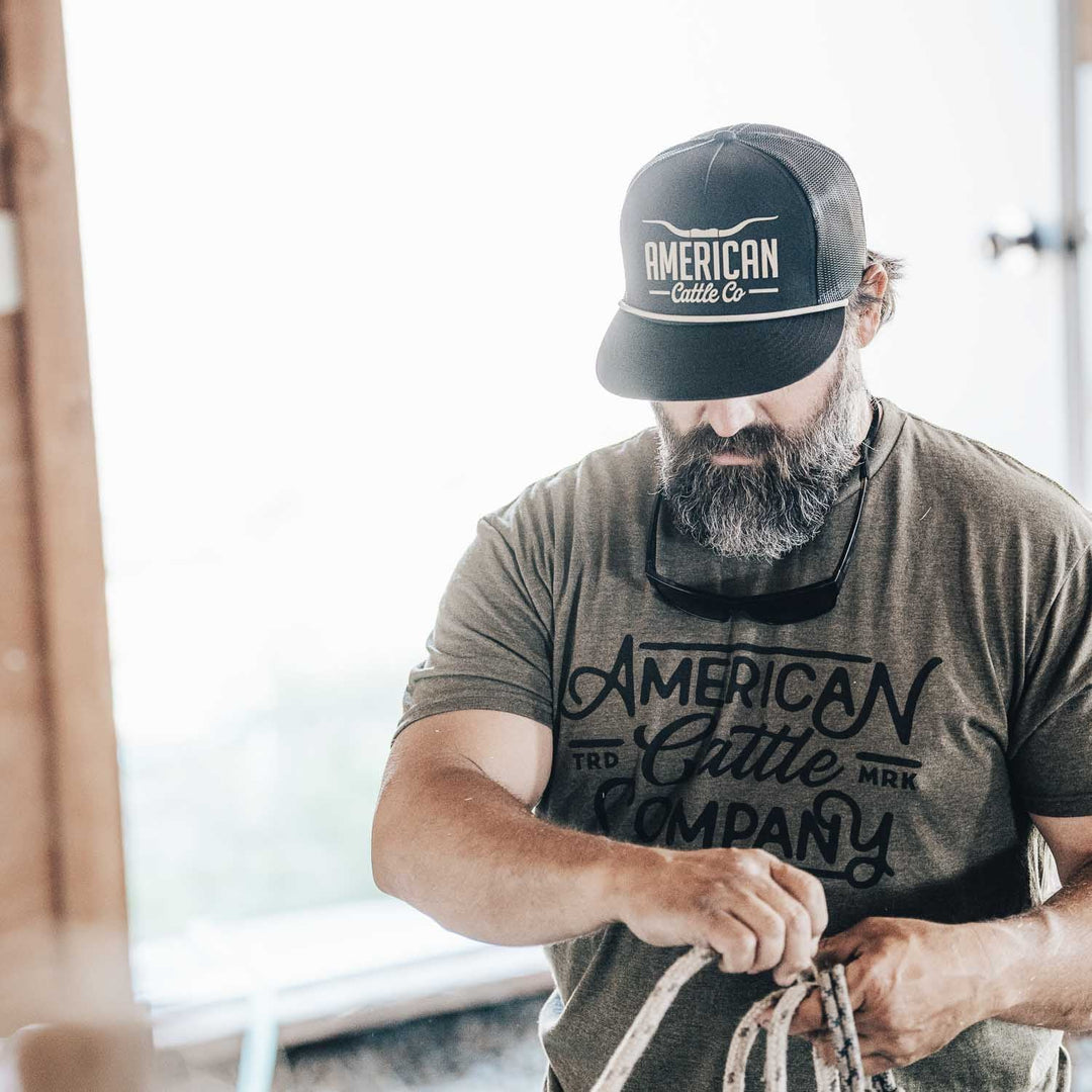 A bearded man in a black "Longhorn Hat" by American Cattle Co and a green t-shirt works with a rope indoors, exemplifying the authentic ranching lifestyle.
