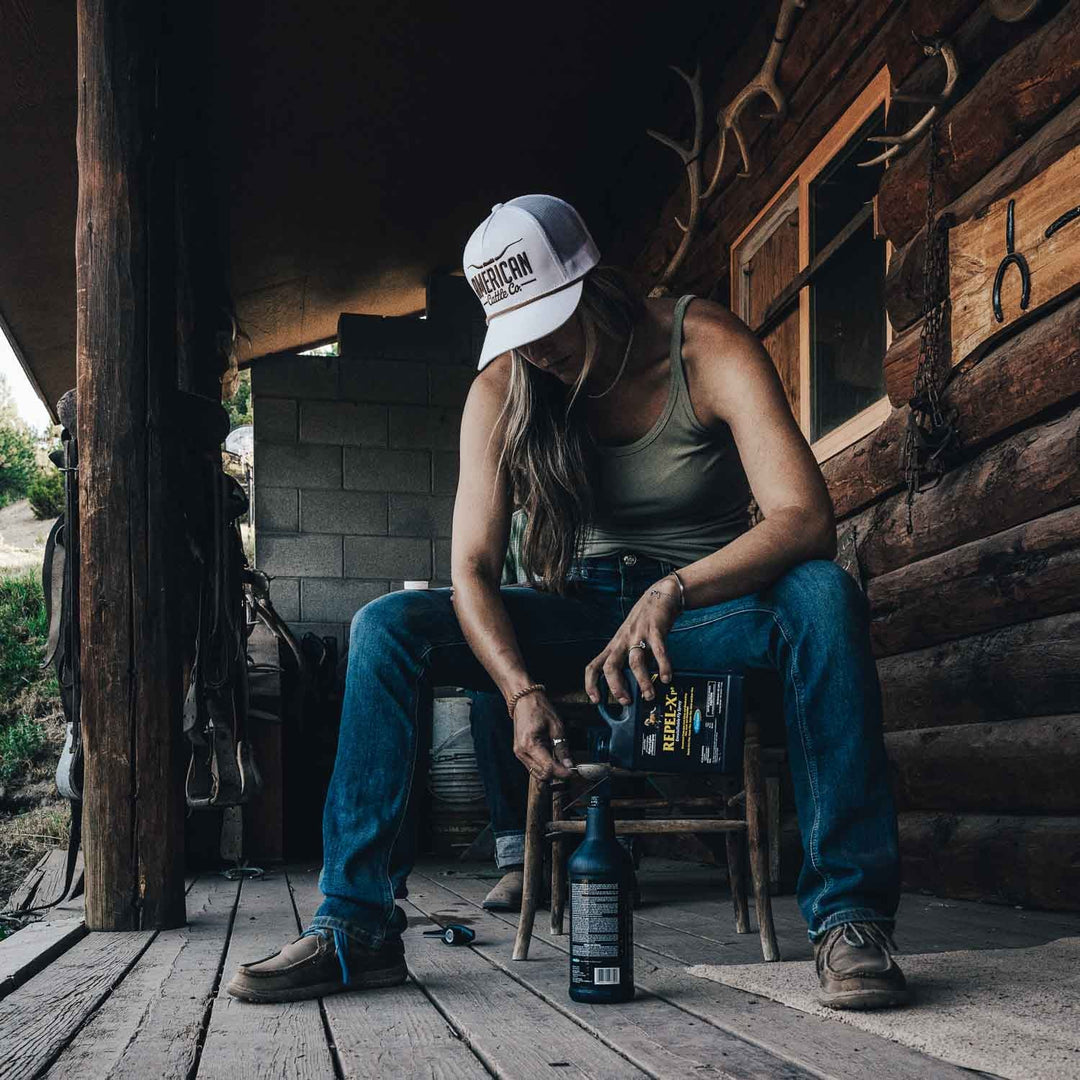 Wearing a white American Cattle Co Longhorn Hat, a tank top, and jeans, a person sits on a wooden porch with a bottle and funnel. A log cabin wall and ranch tools complete the laid-back ranching scene.