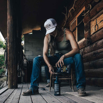 Wearing a white American Cattle Co Longhorn Hat, a tank top, and jeans, a person sits on a wooden porch with a bottle and funnel. A log cabin wall and ranch tools complete the laid-back ranching scene.