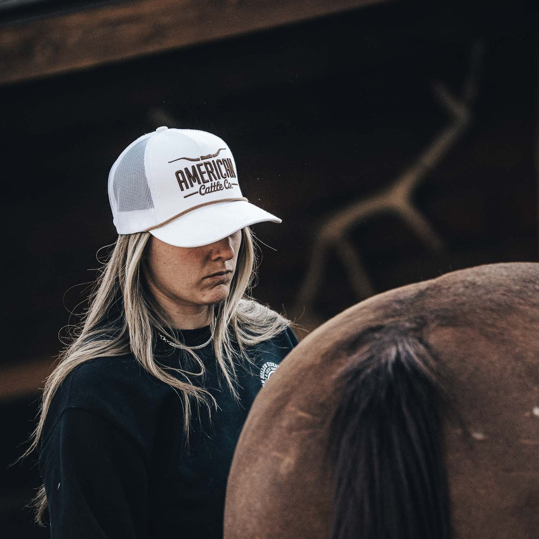 A person with long blonde hair, wearing a white American Cattle Co Longhorn Hat and a black shirt, stands beside a brown horse, embracing the ranching lifestyle with their face partially hidden by the cap.