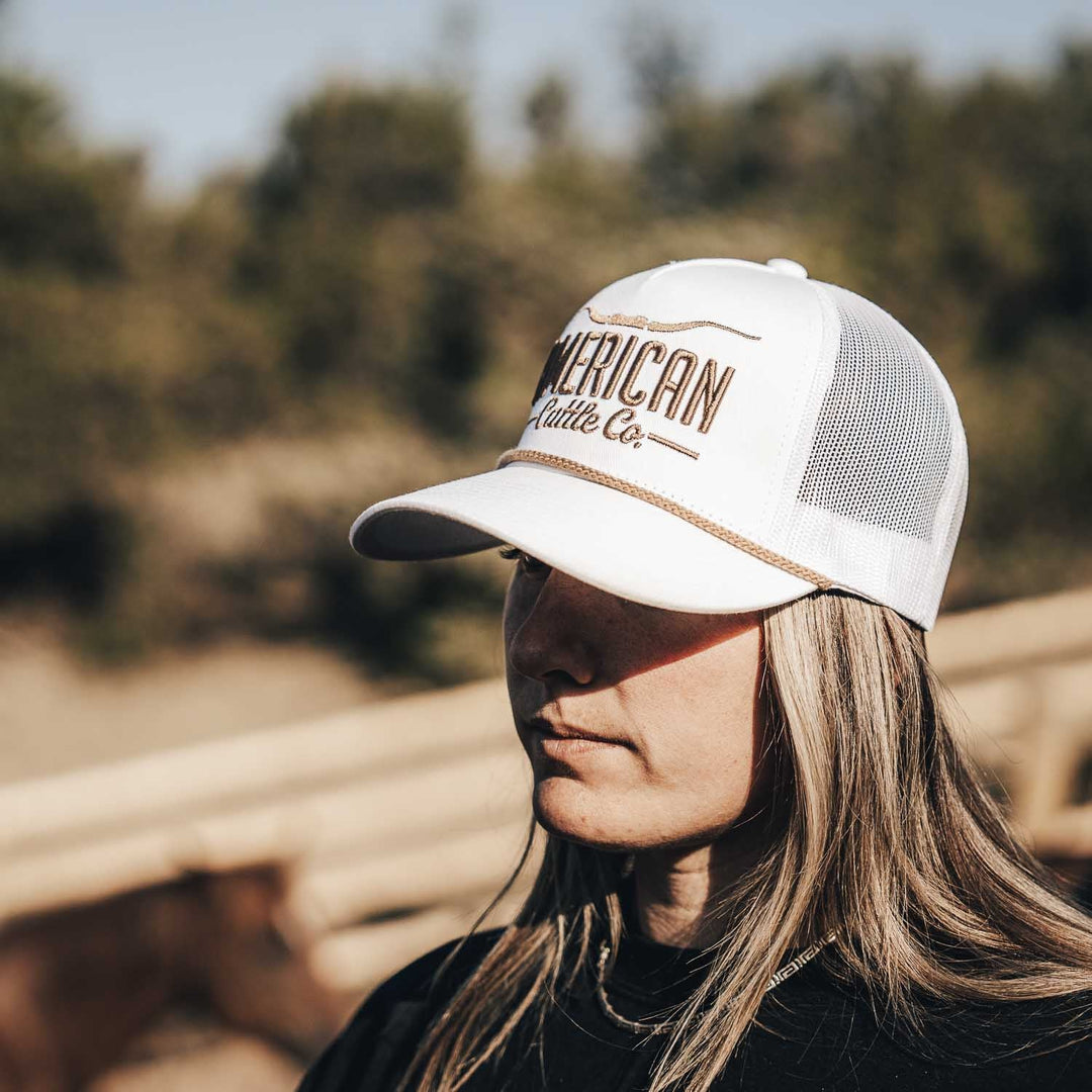 A person with long, light brown hair wears a white Longhorn Hat by American Cattle Co, standing outside near a wooden fence and horse, embracing the ranching lifestyle with blurred trees in the background.