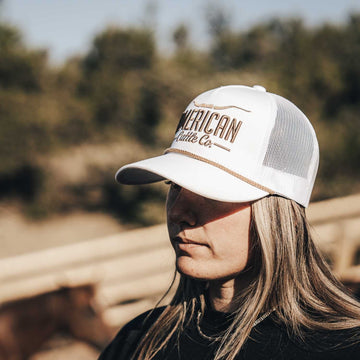A person with long, light brown hair wears a white Longhorn Hat by American Cattle Co, standing outside near a wooden fence and horse, embracing the ranching lifestyle with blurred trees in the background.