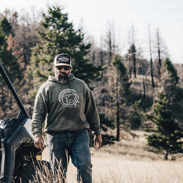 A bearded man in sunglasses, a green hoodie, jeans, and an American Cattle Co Longhorn Hat walks through grass near a parked off-road vehicle, with trees and a hill behind him, embodying the ranching lifestyle.