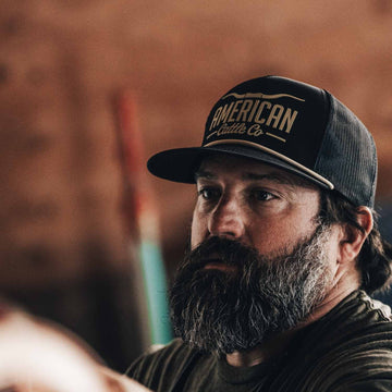 A man with a thick gray beard wears the black Longhorn Hat by American Cattle Co. The blurred, brown-toned background with indistinct shapes suggests his ranching lifestyle.