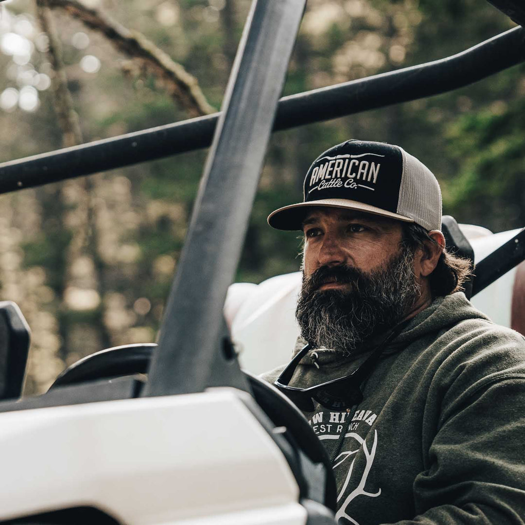 A man with a full beard and mustache, wearing a dark hoodie and the American Cattle Co Longhorn Hat, sits behind the wheel of an off-road vehicle in a wooded outdoor setting, embracing the ranching lifestyle.