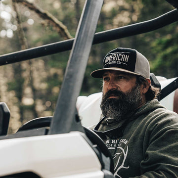 A man with a full beard and mustache, wearing a dark hoodie and the American Cattle Co Longhorn Hat, sits behind the wheel of an off-road vehicle in a wooded outdoor setting, embracing the ranching lifestyle.