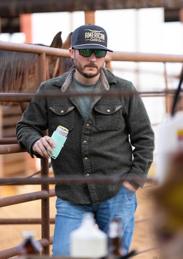 A man in sunglasses and a dark jacket wears the American Cattle Co Longhorn Hat while holding a canned drink by a metal fence, as horses graze nearby and blurred ranching scenery fills the background.