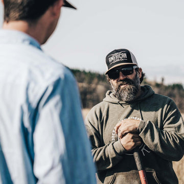 Wearing an American Cattle Co Longhorn Hat, sunglasses, and a hoodie, a man holds a shovel as he faces someone in a light blue shirt outdoors. Trees and clear sky evoke the ranching lifestyle.