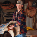 A man in a Rural Cloth Lumberjacket and baseball cap holds a saddle blanket and saddle indoors, with blankets and gear in the background.