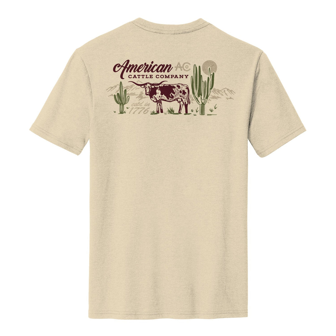 The On the Range Tee by American Cattle Co is a cream poly-cotton shirt with a back graphic of a cow, cacti, and mountains. "American Cattle Company" appears in stylized text above the cow, giving this tee a unique Southwest vibe.