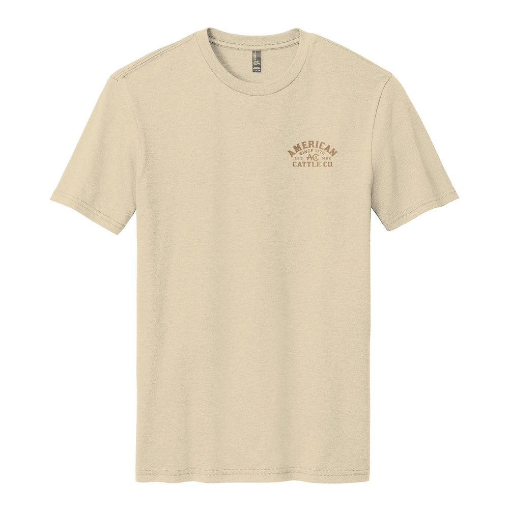 The On the Range Tee by American Cattle Co is a beige poly-cotton short-sleeve shirt with a small brown "AMERICAN CATTLE CO." logo on the left chest, shown laid flat on a white background.