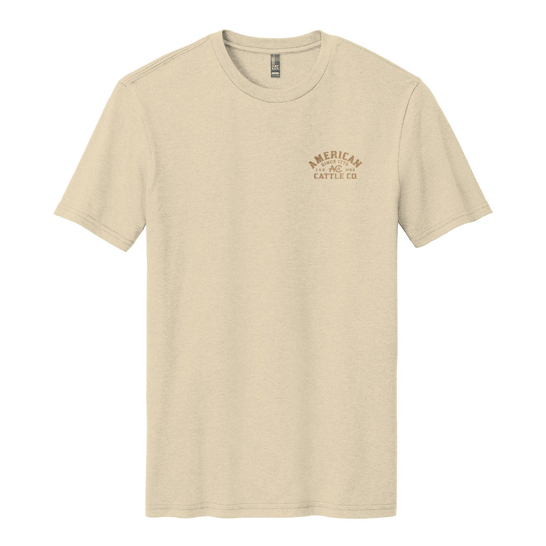 The On the Range Tee by American Cattle Co is a beige poly-cotton short-sleeve shirt with a small brown "AMERICAN CATTLE CO." logo on the left chest, shown laid flat on a white background.