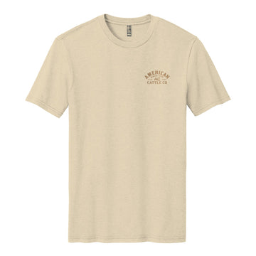 The On the Range Tee by American Cattle Co is a beige poly-cotton short-sleeve shirt with a small brown "AMERICAN CATTLE CO." logo on the left chest, shown laid flat on a white background.