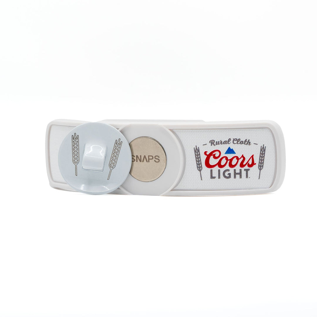 The RC x CL SNAPS Bottle Opener by Rural Cloth is a white belt buckle with a round "SNAPS" snap, silver latch, magnetic bottle opener, and wheat graphics with "Rural Cloth Coors Light" in red and blue.