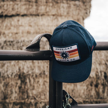 A Retro Spur Hat—a navy blue snapback featuring an “American Cattle Co.” patch—rests on a metal fence, with large hay bales stacked in the blurred background.