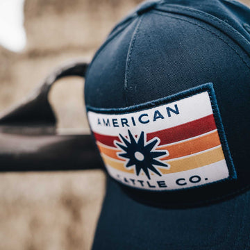 Introducing the Retro Spur Hat—a navy blue snapback ranch hat featuring a rectangular patch that reads “AMERICAN CATTLE CO.” with a stylized sunburst and horizontal stripes in red, orange, and yellow. This vintage-inspired western cap blends classic cowboy ball cap style with bold, retro detailing.