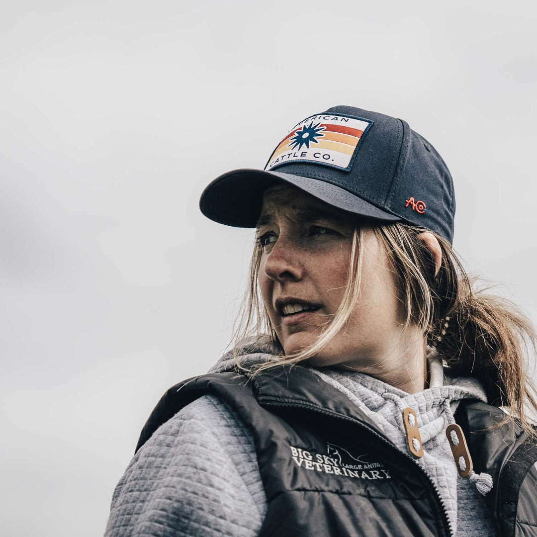 Wearing the Retro Spur Hat with a dark design and logo, a person in a “Big Sky Veterinary” jacket looks off to the side beneath a cloudy sky.